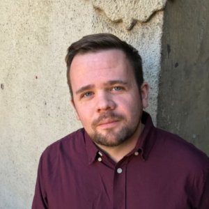 Professional headshot portrait of bearded man in burgundy button-up shirt against textured wall background